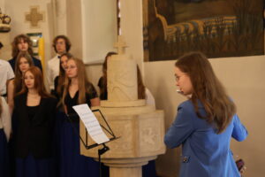 Wnętrze kościoła. Dziewczyna w niebieskiej marynarce gra na flecie poprzecznym, stojąc przed pulpitem z nutami. W tle chór szkolny – uczniowie ubrani w białe koszule i długie niebieskie spódnice lub czarne spodnie. Za nimi znajduje się chrzcielnica wykonana z jasnego kamienia i ściany ozdobione religijnymi malowidłami i symbolami.