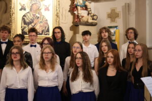 Szkolny chór śpiewa w kościele. Uczniowie stoją w dwóch rzędach, ubrani odświętnie – dziewczęta w białych bluzkach i niebieskich spódnicach, chłopcy w koszulach, część w muszkach. W tle widoczne są rzeźby i duży obraz Matki Boskiej Częstochowskiej z Dzieciątkiem, otoczony złotą ramą. Atmosfera poważna i uroczysta.