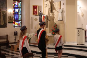 Trójka uczniów w poczcie sztandarowym w kościele. Dziewczyny mają białe koszule i ciemne spódnice, chłopak – garnitur. Wszyscy mają na głowach granatowe czapki z czerwonymi elementami i czerwone szarfy. Stoją przed ołtarzem z rzeźbami Jana Pawła II i świętych. Po lewej stronie widoczne kolorowe witraże.