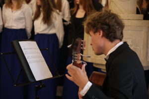 Zbliżenie na chłopaka grającego na gitarze klasycznej. Siedzi zwrócony bokiem, skupiony na nutach ustawionych na czarnym pulpicie. W tle stojący członkowie chóru w białych koszulach i niebieskich spódnicach, patrzący w stronę grającego. Scena rozgrywa się w kościele.