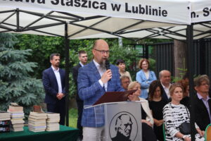 Mężczyzna z długą brodą, w niebieskiej marynarce w kratę, przemawia do mikrofonu stojąc za mównicą z logo szkoły. W lewej dłoni trzyma dokument, przed nim na stole leżą stosy książek. W tle osoby siedzące i stojące – nauczyciele i zaproszeni goście, z powagą słuchający wystąpienia.