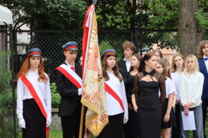 Grupa uczniów i poczet sztandarowy stoi na zewnątrz, obok siatki i drzewa. Troje uczniów w poczcie trzyma sztandar szkoły – na czerwono-białej tkaninie widać datę „1884” i nazwę szkoły. Młodzież ubrana jest elegancko, przeważnie w białe koszule i czarne spodnie lub spódnice. Dziewczyna z lewej strony ma długie rude włosy i poważny wyraz twarzy.