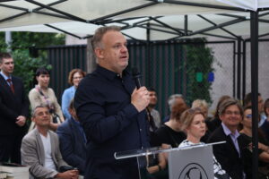 Mężczyzna w granatowej koszuli z mikrofonem przemawia do zgromadzonych. Stoi przy przezroczystej mównicy z logo szkoły – profilem Stanisława Staszica. W tle publiczność siedzi na zielonych krzesłach – kobiety i mężczyźni w eleganckich strojach, część uśmiechnięta, część skupiona.