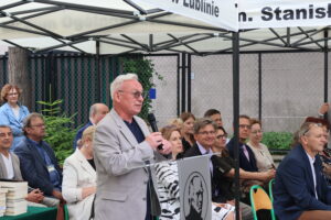 Starszy mężczyzna z siwymi włosami i okularami, ubrany w jasnoszarą marynarkę w kratkę i ciemny T-shirt, przemawia z mikrofonem. Stoi przy mównicy z logo szkoły. W tle rzędy siedzących gości – nauczycieli i zaproszonych – w eleganckich strojach, z zainteresowaniem słuchających. Namiot z napisem „I Liceum Ogólnokształcące im. Stanisława Staszica w Lublinie”.