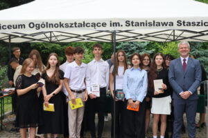 Grupa 13 uczniów – 6 chłopców i 7 dziewczyn – pozuje do zdjęcia pamiątkowego. Stoją przed namiotem szkoły. Wszyscy w galowych strojach – białe koszule, czarne sukienki, spódnice, spodnie. Niektórzy trzymają książki, m.in. „Nic mnie nie powstrzyma” i „Dante”. Obok nich uśmiechnięty nauczyciel w granatowym garniturze i czerwonym krawacie.