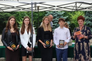Czworo uczniów – trzy dziewczyny i chłopak – oraz nauczycielka pozują z książkami. Dziewczyny w eleganckich sukienkach i marynarkach – dwie trzymają książki z serii „Igrzyska śmierci”. Chłopak w białej koszuli i ciemnych spodniach trzyma „Milczenie owiec” Thomasa Harrisa. Nauczycielka w granatowej sukni z jasnym, roślinnym wzorem trzyma książkę ks. Tischnera.