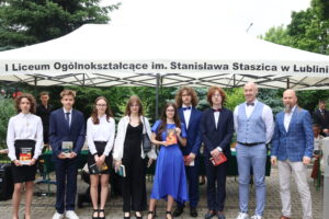 Dziewięcioro uczniów (4 dziewczyny, 5 chłopców) pozuje pod namiotem z nauczycielami. Chłopcy w garniturach, dziewczyny w białych bluzkach, jedna w niebieskiej sukni. Trzymają książki różnych autorów. Obok nich dwóch nauczycieli – jeden w jasnoniebieskiej kamizelce i muszce, drugi z długą brodą i w kratkowanej marynarce.