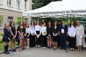 Grupa 13 uczniów – głównie dziewczyny – pozuje z nauczycielką w jasnej sukience lnianej. Uczennice w czerni i bieli – spódnice, marynarki, koszule; część trzyma książki. Budynek szkoły widoczny w tle – jasna elewacja, otwarte okna, ozdobne krzewy. Namiot z nazwą szkoły na górze kadru.