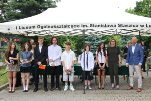 Dziewięcioro uczniów – czterech chłopców i pięć dziewcząt – pozuje pod namiotem. Dziewczęta w sukienkach, spódnicach i bluzkach; chłopcy w koszulach i garniturach. Jeden chłopiec ma dłuższe włosy i białą, luźną koszulę. Obok nauczycielka w zielonym kombinezonie i nauczyciel z brodą w niebieskiej marynarce.