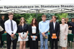 Sześciu uczniów (3 dziewczyny, 3 chłopców) stoi w rzędzie z książkami w dłoniach. Wszyscy ubrani elegancko – dziewczyny w ciemnych spódnicach i bluzkach, chłopcy w koszulach, jeden w krawacie. Obok nauczycielka w kremowym żakiecie i okularach. W tle namiot szkoły i stół z książkami.