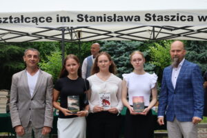 Na tle białego namiotu z napisem „I Liceum Ogólnokształcące im. Stanisława Staszica w Lublinie” stoi pięć osób. Po bokach dwaj nauczyciele – po lewej mężczyzna w jasnoszarej marynarce, po prawej – mężczyzna z długą brodą w niebieskiej marynarce w kratę. Pośrodku stoją trzy uczennice – wszystkie trzymają książki. Pierwsza z lewej ma czarną bluzkę i jasną spódnicę, trzyma książkę pt. „Schronisko” Remigiusza Mroza. Druga uczennica ubrana jest w białą koszulę i czarne spodnie, trzyma książkę Jo Nesbø pt. „Wybawiciel”. Trzecia uczennica, w białej bluzce z falbankami, trzyma książkę „Schronisko. Tom 2: Przetrwanie”. W tle widać mężczyznę w kamizelce i krawacie oraz drzewa iglaste.