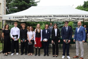 Dziewięć osób pozuje wspólnie pod namiotem z nazwą szkoły. Po lewej stronie stoi kobieta w długiej, czarnej sukience – nauczycielka. Obok niej siedmioro uczniów – czterech chłopców i trzy dziewczęta. Uczniowie ubrani są galowo – białe koszule, czarne spodnie lub spódnice, jeden chłopiec w garniturze i muszce. Trzymają książki – wśród nich „Michio Kaku: Fizyka rzeczy niemożliwych”, „Toskania”, „Futu.re” Glukhovsky’ego. Po prawej stronie stoi nauczyciel w niebieskiej marynarce w kratę. Tło stanowi zieleń i sosnowe drzewa.