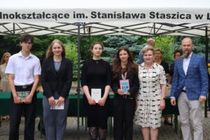 Sześć osób stoi w rzędzie – czterech uczniów i dwójka nauczycieli. Po lewej chłopak w białej koszuli i czarnych spodniach, trzyma książkę pt. „Kiedy góry płaczą” Greena. Obok trzy dziewczyny w eleganckich strojach – czarne sukienki, marynarki. Jedna z nich trzyma książkę z biało-niebieską okładką, druga – kolorową książkę z rysunkiem budynku na okładce. Po prawej stronie nauczycielka w sukience w geometryczny wzór i nauczyciel w niebieskiej marynarce. W tle uczniowie i zielone drzewa.