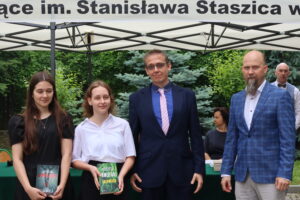Czwórka osób stoi pod namiotem – dwie uczennice i dwóch nauczycieli. Po lewej uczennica z długimi, ciemnymi włosami w czarnej sukience trzyma książkę „W pułapce” Magdy Stachuli. Obok druga uczennica w białej bluzce i czarnej spódnicy trzyma książkę „Zmułowisko” Wojciecha Chmielarza. Nauczyciel w środku ma granatowy garnitur i różowo-białą krawatkę. Po prawej nauczyciel w niebieskiej marynarce w kratę. W tle sosny i krzewy ozdobne.