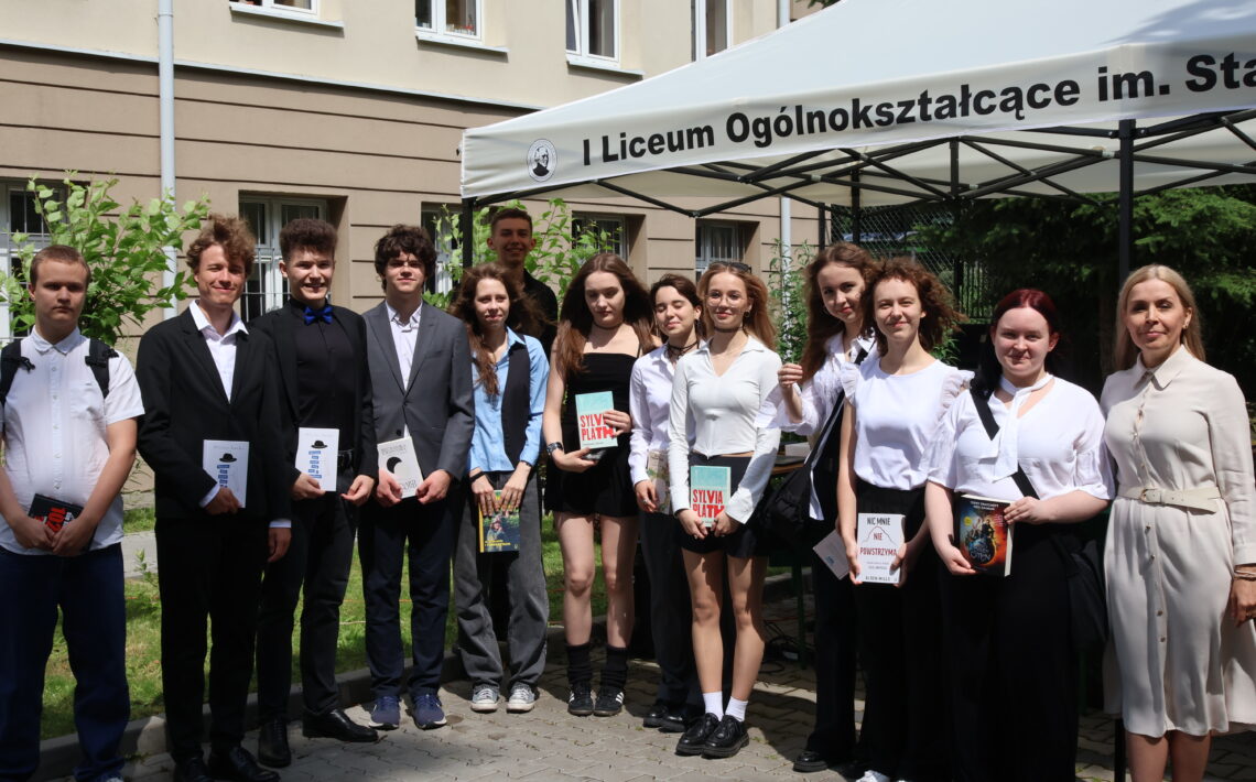 Grupa 15 uczniów i nauczycielka po prawej stronie stoją w pełnym słońcu, przed namiotem i budynkiem szkoły. Uczniowie – dziewięć dziewcząt i sześciu chłopców – ubrani są elegancko, w różnorodne stylizacje galowe: marynarki, koszule, sukienki, spódnice. Dziewczęta trzymają książki, m.in. „Szklany klosz” Sylvii Plath, „Nic mnie nie powstrzyma” Adama Smitha oraz powieści fantasy. Nauczycielka z prawej strony ubrana w kremową sukienkę z paskiem. W tle okna szkoły i ozdobna roślinność.