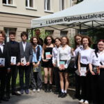 Grupa 15 uczniów i nauczycielka po prawej stronie stoją w pełnym słońcu, przed namiotem i budynkiem szkoły. Uczniowie – dziewięć dziewcząt i sześciu chłopców – ubrani są elegancko, w różnorodne stylizacje galowe: marynarki, koszule, sukienki, spódnice. Dziewczęta trzymają książki, m.in. „Szklany klosz” Sylvii Plath, „Nic mnie nie powstrzyma” Adama Smitha oraz powieści fantasy. Nauczycielka z prawej strony ubrana w kremową sukienkę z paskiem. W tle okna szkoły i ozdobna roślinność.