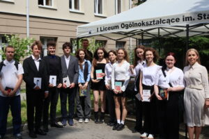 Grupa 15 uczniów i nauczycielka po prawej stronie stoją w pełnym słońcu, przed namiotem i budynkiem szkoły. Uczniowie – dziewięć dziewcząt i sześciu chłopców – ubrani są elegancko, w różnorodne stylizacje galowe: marynarki, koszule, sukienki, spódnice. Dziewczęta trzymają książki, m.in. „Szklany klosz” Sylvii Plath, „Nic mnie nie powstrzyma” Adama Smitha oraz powieści fantasy. Nauczycielka z prawej strony ubrana w kremową sukienkę z paskiem. W tle okna szkoły i ozdobna roślinność.