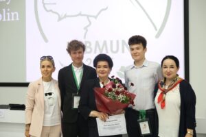 Pięć osób stoi w szeregu na tle białej ściany z dużym, wyświetlonym logo „LUBMUN 2024”. W centrum starsza kobieta z elegancką, ciemną fryzurą trzyma bukiet czerwonych i białych kwiatów oraz dyplom z podziękowaniami – to najprawdopodobniej osoba nagradzana. Po jej lewej stronie stoi młody mężczyzna w garniturze z zieloną smyczą i identyfikatorem. Z prawej strony starszej kobiety jest drugi młody mężczyzna w jasnej koszuli z identyfikatorem na zielonej smyczy. Na skrajach stoją dwie kobiety: po lewej blondynka w jasnym garniturze i białym t-shircie z grafiką kota w okularach przeciwsłonecznych, po prawej brunetka z kolorową apaszką. Wszyscy uśmiechają się i patrzą w stronę obiektywu. W tle widać fragment białego ekranu z elementami logo.