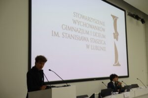 Zdjęcie przedstawia młodego mężczyznę stojącego przy mównicy. Ma jasnobrązowe włosy, jest ubrany na czarno i mówi do mikrofonu. Po prawej stronie kadru, przy stole prezydialnym, siedzi druga osoba – młody mężczyzna z kręconymi włosami i zieloną smyczą – także przemawia. Tło to duży biały ekran z brązowym napisem: „STOWARZYSZENIE WYCHOWANKÓW GIMNAZJUM I LICEUM IM. STANISŁAWA STASZICA W LUBLINIE” oraz stylizowaną literą „I” z liściem winorośli.