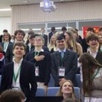 Zdjęcie ukazuje tłum uczniów stojących w auli. Są to głównie nastolatkowie, ubrani w eleganckie stroje – marynarki, koszule, krawaty. Wszyscy mają na szyjach zielone smycze z identyfikatorami. Wśród nich widoczna jest dziewczyna owinięta flagą Wielkiej Brytanii. Na środku kadru chłopak z ręką na piersi – gest oddający hołd lub szacunek. W tle nad ich głowami widać dużą, rozciągniętą flagę Chin. Wszyscy są uśmiechnięci i wyraźnie zaangażowani w wydarzenie.