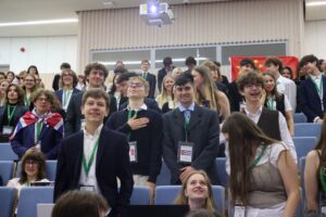 Zdjęcie ukazuje tłum uczniów stojących w auli. Są to głównie nastolatkowie, ubrani w eleganckie stroje – marynarki, koszule, krawaty. Wszyscy mają na szyjach zielone smycze z identyfikatorami. Wśród nich widoczna jest dziewczyna owinięta flagą Wielkiej Brytanii. Na środku kadru chłopak z ręką na piersi – gest oddający hołd lub szacunek. W tle nad ich głowami widać dużą, rozciągniętą flagę Chin. Wszyscy są uśmiechnięci i wyraźnie zaangażowani w wydarzenie.