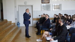 Wystąpienie oficjalne, najpewniej inauguracyjne. Starszy mężczyzna w garniturze i okularach, trzymający mikrofon, przemawia do widowni z lewej strony sali. W tle na ścianie widnieją marmurowe tablice pamiątkowe i cytat łaciński „Salus rei publicae suprema lex esto”. Mężczyzna kieruje się w stronę pierwszego rzędu, gdzie siedzą dorośli, m.in. mężczyzna w zielonym golfie i drugi w ciemnym garniturze. Dalej – liczni młodzi uczestnicy, głównie młodzież.