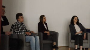 Cztery młode osoby siedzą na wygodnych fotelach ustawionych w półkolu, najprawdopodobniej panel dyskusyjny. Dwie kobiety i dwóch mężczyzn. Osoby są ubrane elegancko, ale swobodnie – jedna z kobiet ma granatową marynarkę i białą bluzkę koronkową, druga bordową marynarkę. Jeden z mężczyzn ma koszulę w kratę i jasne dżinsy. Drugi, częściowo widoczny, trzyma mikrofon. W tle – pusta ściana.