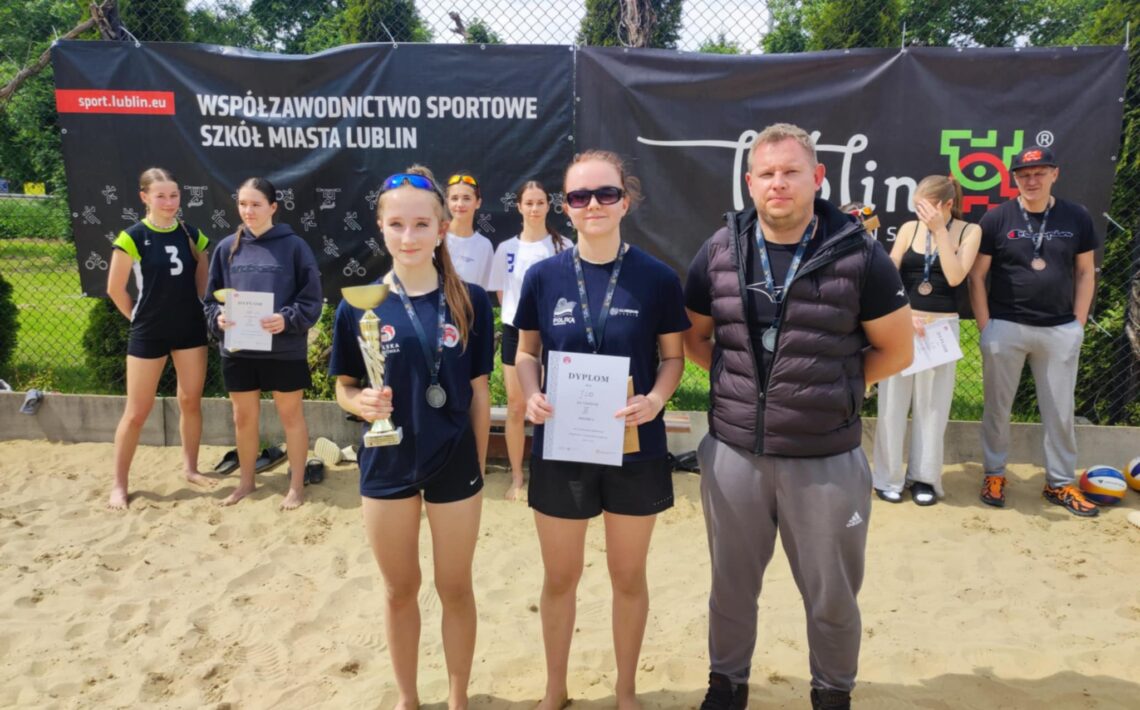 Grupa uczestników – boisko do siatkówki plażowej na zewnątrz: Tło: Za uczestnikami znajduje się czarne ogrodzenie z banerami reklamowymi. Jeden z banerów to czarny prostokąt z białym napisem „Współzawodnictwo sportowe szkół miasta Lublin”, z logo „sport.lublin.eu”. Obok drugi baner z napisem „Lublin”, logo miasta Lublin z kolorowym, geometrycznym symbolem. Uczestnicy: Wszyscy ubrani w sportowe stroje. Dziewczęta mają na sobie czarne lub granatowe koszulki sportowe i krótkie spodenki. W tle widać również kilka osób w białych koszulkach, jedna z nich trzyma piłkę do siatkówki. Pierwszy plan: Dwie młode dziewczyny stoją z przodu, trzymają trofea – jedna z nich puchar złoty, druga trzyma dyplom w białej kartce formatu A4 z dużym napisem „DYPLOM”. Obie dziewczyny mają ciemne koszulki i sportowe medale na szyjach. Obok nich stoi mężczyzna w czarnej kamizelce, szare spodnie dresowe, czarne buty sportowe – prawdopodobnie trener lub opiekun. Ma krótko ścięte włosy, ręce założone z tyłu. Pozostałe osoby w tle: Kilka dziewczyn, niektóre w koszulkach z numerami (np. numer 3), trzymają dyplomy lub stoją z medalami na szyjach. Część uczestników ma okulary przeciwsłoneczne. Podłoże: Piasek boiska do siatkówki, gołe stopy uczestników. Atmosfera: Radość i duma, uśmiechy na twarzach, zawodnicy stoją w pozie do pamiątkowego zdjęcia.