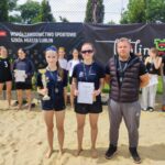 Grupa uczestników – boisko do siatkówki plażowej na zewnątrz: Tło: Za uczestnikami znajduje się czarne ogrodzenie z banerami reklamowymi. Jeden z banerów to czarny prostokąt z białym napisem „Współzawodnictwo sportowe szkół miasta Lublin”, z logo „sport.lublin.eu”. Obok drugi baner z napisem „Lublin”, logo miasta Lublin z kolorowym, geometrycznym symbolem. Uczestnicy: Wszyscy ubrani w sportowe stroje. Dziewczęta mają na sobie czarne lub granatowe koszulki sportowe i krótkie spodenki. W tle widać również kilka osób w białych koszulkach, jedna z nich trzyma piłkę do siatkówki. Pierwszy plan: Dwie młode dziewczyny stoją z przodu, trzymają trofea – jedna z nich puchar złoty, druga trzyma dyplom w białej kartce formatu A4 z dużym napisem „DYPLOM”. Obie dziewczyny mają ciemne koszulki i sportowe medale na szyjach. Obok nich stoi mężczyzna w czarnej kamizelce, szare spodnie dresowe, czarne buty sportowe – prawdopodobnie trener lub opiekun. Ma krótko ścięte włosy, ręce założone z tyłu. Pozostałe osoby w tle: Kilka dziewczyn, niektóre w koszulkach z numerami (np. numer 3), trzymają dyplomy lub stoją z medalami na szyjach. Część uczestników ma okulary przeciwsłoneczne. Podłoże: Piasek boiska do siatkówki, gołe stopy uczestników. Atmosfera: Radość i duma, uśmiechy na twarzach, zawodnicy stoją w pozie do pamiątkowego zdjęcia.