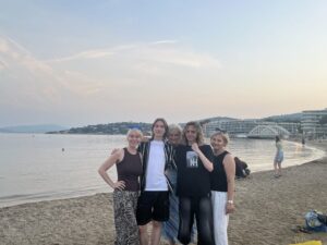 Zdjęcie wykonane wieczorem na piaszczystej plaży. W tle spokojne morze, budynki hotelowe i charakterystyczny biały mostek nad wodą. Na pierwszym planie stoi pięć osób – trzy kobiety i dwóch młodych mężczyzn. Wszyscy się uśmiechają. Kobieta po lewej ma fioletowe okulary przeciwsłoneczne na głowie i czarną sukienkę, środkowy chłopak ma na sobie biały t-shirt i rozpiętą czarno-białą koszulę w paski, druga dziewczyna ma czarne spodnie i czarną koszulkę z logo NH, kobieta po prawej – czarną bluzkę bez rękawów i jasne spodnie w paski. Osoby stoją blisko siebie z objętymi ramionami.