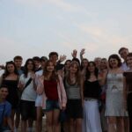 Zdjęcie grupowe wykonane na plaży, o zmierzchu. Na tle spokojnego morza i wieczornego nieba stoi kilkanaście młodych osób (zarówno dziewczyny, jak i chłopcy), ustawionych w kilku rzędach, z rękami podniesionymi w geście powitania. Osoby ubrane są w letnie stroje – sukienki, topy, szorty. W tle widać wzgórza i rozświetlone niebo z resztką dziennego światła. Atmosfera jest radosna i przyjazna.
