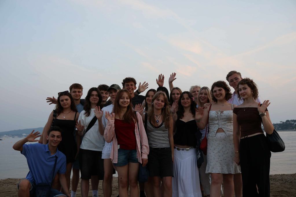 Zdjęcie grupowe wykonane na plaży, o zmierzchu. Na tle spokojnego morza i wieczornego nieba stoi kilkanaście młodych osób (zarówno dziewczyny, jak i chłopcy), ustawionych w kilku rzędach, z rękami podniesionymi w geście powitania. Osoby ubrane są w letnie stroje – sukienki, topy, szorty. W tle widać wzgórza i rozświetlone niebo z resztką dziennego światła. Atmosfera jest radosna i przyjazna.