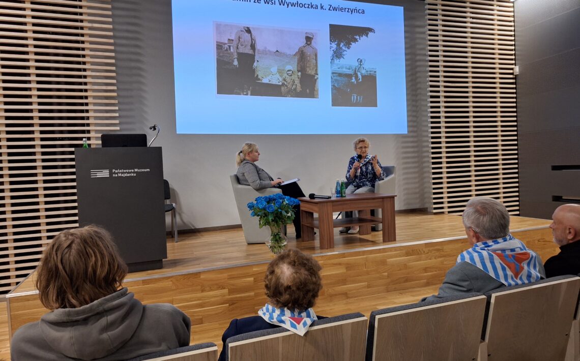 Zdjęcie przedstawia wydarzenie odbywające się w nowoczesnej sali konferencyjnej z drewnianą podłogą i ścianami o nowoczesnym wykończeniu. Na podwyższeniu na scenie znajdują się dwie kobiety siedzące w fotelach. Kobieta po lewej stronie ma blond włosy spięte w kucyk i ubrana jest w szary strój. Kobieta po prawej stronie, prowadząca rozmowę, ma jasne włosy i kolorową bluzkę z wzorami. Mówi do mikrofonu i gestykuluje rękami. Przed nią stoi drewniany stolik z dwiema butelkami wody i mikrofonem. Obok stolika postawiony jest bukiet niebieskich róż w przezroczystym wazonie — te same kwiaty co na poprzednim zdjęciu. Na ekranie za prelegentkami znajduje się biały slajd z niebieskim tytułem: „Rodzina Wolamin ze wsi Wywłoczka k. Zwierzyńca”. Poniżej widoczne są dwa czarno-białe zdjęcia archiwalne przedstawiające ludzi na tle wiejskiego krajobrazu. Na pierwszym planie, na widowni, siedzi kilka osób. Niektóre z nich mają na plecach biało-niebieskie szaliki w pasy z czerwonym symbolem — są to najprawdopodobniej elementy symboliki historycznej lub pamiątkowej, np. obozowej. Wśród słuchaczy można rozpoznać chłopca z pierwszego zdjęcia — ma tę samą fryzurę i szarą bluzę z kapturem. Na lewej stronie sceny znajduje się ciemna mównica z białym napisem: „Państwowe Muzeum na Majdanku”.