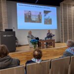 Zdjęcie przedstawia wydarzenie odbywające się w nowoczesnej sali konferencyjnej z drewnianą podłogą i ścianami o nowoczesnym wykończeniu. Na podwyższeniu na scenie znajdują się dwie kobiety siedzące w fotelach. Kobieta po lewej stronie ma blond włosy spięte w kucyk i ubrana jest w szary strój. Kobieta po prawej stronie, prowadząca rozmowę, ma jasne włosy i kolorową bluzkę z wzorami. Mówi do mikrofonu i gestykuluje rękami. Przed nią stoi drewniany stolik z dwiema butelkami wody i mikrofonem. Obok stolika postawiony jest bukiet niebieskich róż w przezroczystym wazonie — te same kwiaty co na poprzednim zdjęciu. Na ekranie za prelegentkami znajduje się biały slajd z niebieskim tytułem: „Rodzina Wolamin ze wsi Wywłoczka k. Zwierzyńca”. Poniżej widoczne są dwa czarno-białe zdjęcia archiwalne przedstawiające ludzi na tle wiejskiego krajobrazu. Na pierwszym planie, na widowni, siedzi kilka osób. Niektóre z nich mają na plecach biało-niebieskie szaliki w pasy z czerwonym symbolem — są to najprawdopodobniej elementy symboliki historycznej lub pamiątkowej, np. obozowej. Wśród słuchaczy można rozpoznać chłopca z pierwszego zdjęcia — ma tę samą fryzurę i szarą bluzę z kapturem. Na lewej stronie sceny znajduje się ciemna mównica z białym napisem: „Państwowe Muzeum na Majdanku”.