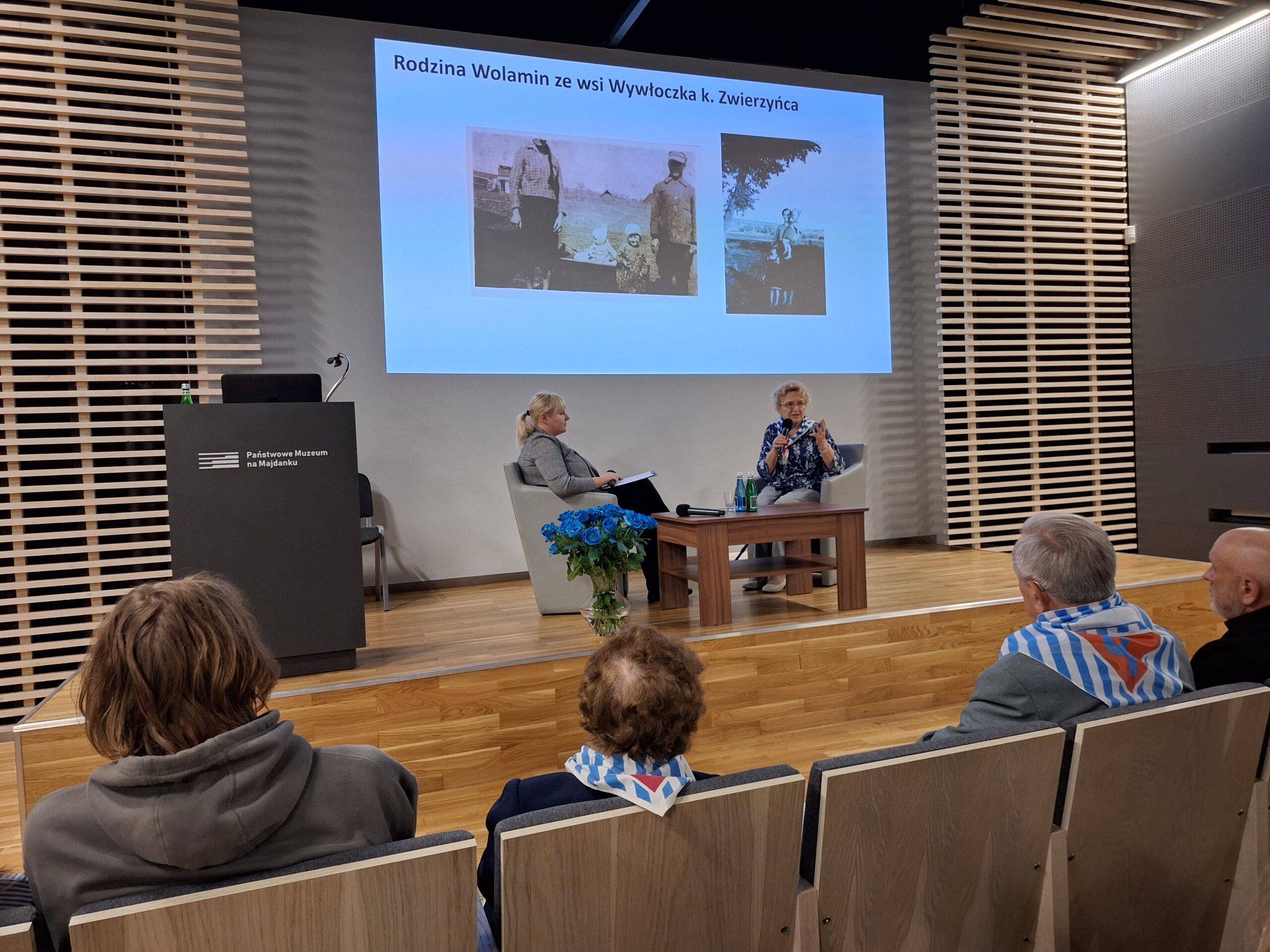 Zdjęcie przedstawia wydarzenie odbywające się w nowoczesnej sali konferencyjnej z drewnianą podłogą i ścianami o nowoczesnym wykończeniu. Na podwyższeniu na scenie znajdują się dwie kobiety siedzące w fotelach. Kobieta po lewej stronie ma blond włosy spięte w kucyk i ubrana jest w szary strój. Kobieta po prawej stronie, prowadząca rozmowę, ma jasne włosy i kolorową bluzkę z wzorami. Mówi do mikrofonu i gestykuluje rękami. Przed nią stoi drewniany stolik z dwiema butelkami wody i mikrofonem. Obok stolika postawiony jest bukiet niebieskich róż w przezroczystym wazonie — te same kwiaty co na poprzednim zdjęciu. Na ekranie za prelegentkami znajduje się biały slajd z niebieskim tytułem: „Rodzina Wolamin ze wsi Wywłoczka k. Zwierzyńca”. Poniżej widoczne są dwa czarno-białe zdjęcia archiwalne przedstawiające ludzi na tle wiejskiego krajobrazu. Na pierwszym planie, na widowni, siedzi kilka osób. Niektóre z nich mają na plecach biało-niebieskie szaliki w pasy z czerwonym symbolem — są to najprawdopodobniej elementy symboliki historycznej lub pamiątkowej, np. obozowej. Wśród słuchaczy można rozpoznać chłopca z pierwszego zdjęcia — ma tę samą fryzurę i szarą bluzę z kapturem. Na lewej stronie sceny znajduje się ciemna mównica z białym napisem: „Państwowe Muzeum na Majdanku”.