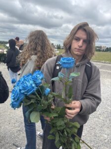 Na pierwszym planie znajduje się młody chłopiec o jasnej cerze i długich, brązowych, rozpuszczonych włosach. Ma poważny wyraz twarzy i patrzy prosto w obiektyw. Ubrany jest w szarą bluzę z kapturem z delikatnym, wytłaczanym napisem oraz ma na plecach czarny plecak. W dłoniach trzyma duży bukiet intensywnie niebieskich róż z zielonymi łodygami i liśćmi — około dziewięciu kwiatów.

Za chłopcem widać grupę kilku osób stojących na utwardzonym żwirowym podłożu, z lewej strony zdjęcia. Osoby są częściowo odwrócone plecami do aparatu. Jedna dziewczyna z długimi, kręconymi blond włosami jest dobrze widoczna z tyłu. Część osób trzyma wieńce z kwiatami w kolorach czerwonym i białym. W tle rozpościera się rozległa, zielona przestrzeń trawiasta. Na horyzoncie widać niskie zabudowania i kilka drzew. Niebo jest całkowicie pokryte ciężkimi, szarymi chmurami — sugeruje pochmurny dzień.