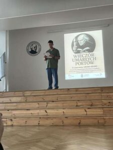 Młody chłopak stoi samotnie na scenie, w półformalnym stroju: zielona koszula i niebieskie spodnie. Trzyma kartkę i czyta. Za nim duży plakat „Wieczoru Umarłych Poetów”. Otoczenie minimalistyczne, drewniane stopnie sceny i biała ściana z logo szkoły.