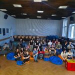 Zdjęcie grupowe wszystkich uczestników wydarzenia. Młodzież w różnym wieku, około 30 osób, siedzi na kolorowych pufach, krzesłach i leżakach w dużej, jasnej sali z drewnianą podłogą i wysokimi oknami. W tle stoły z napojami i przekąskami. Większość ubrana swobodnie, nieformalnie. Część osób pozuje do zdjęcia z uśmiechami, niektórzy robią gesty "peace". Atmosfera bardzo swobodna, integracyjna.