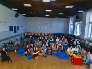 Zdjęcie grupowe wszystkich uczestników wydarzenia. Młodzież w różnym wieku, około 30 osób, siedzi na kolorowych pufach, krzesłach i leżakach w dużej, jasnej sali z drewnianą podłogą i wysokimi oknami. W tle stoły z napojami i przekąskami. Większość ubrana swobodnie, nieformalnie. Część osób pozuje do zdjęcia z uśmiechami, niektórzy robią gesty "peace". Atmosfera bardzo swobodna, integracyjna.