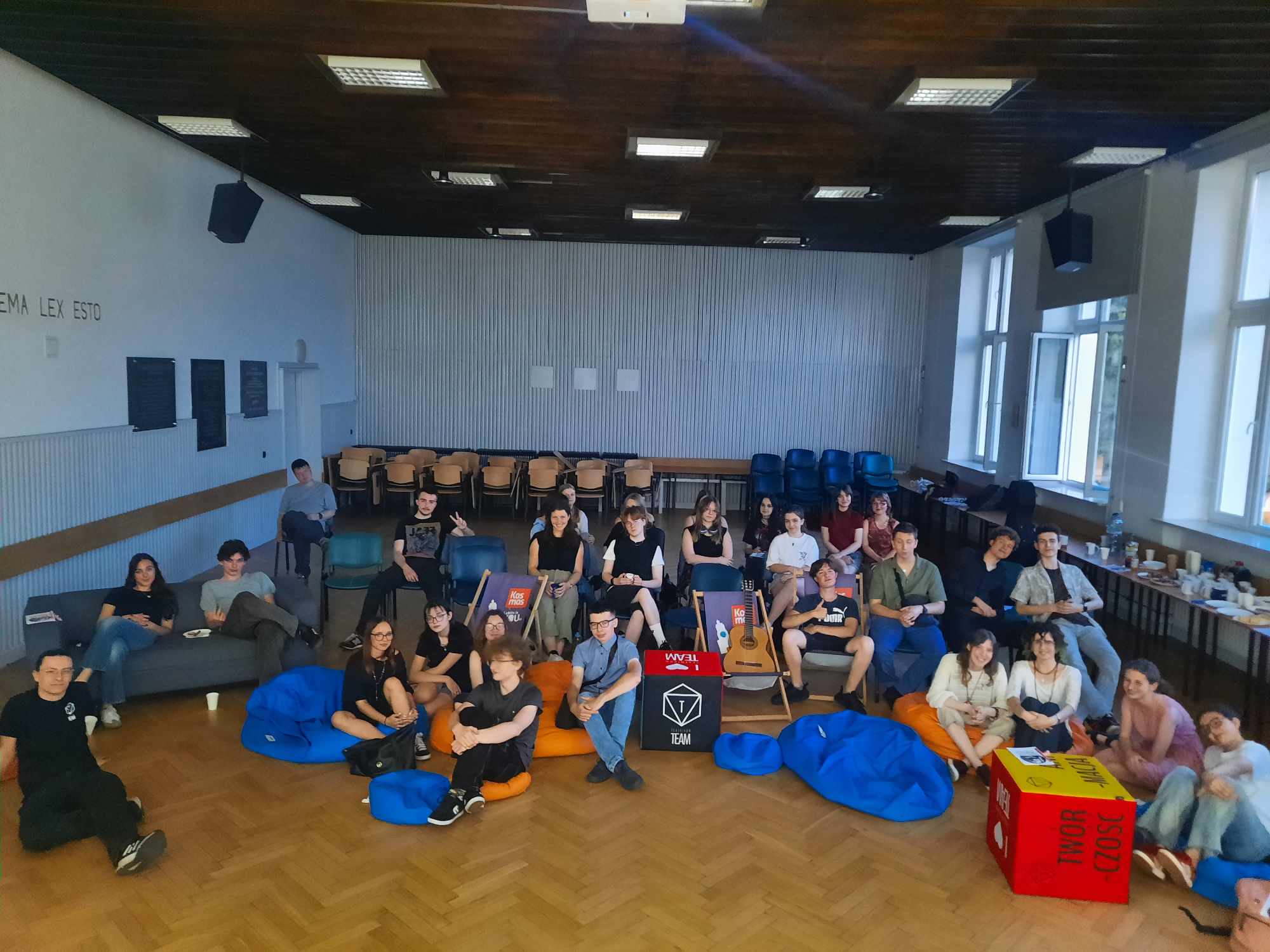 Zdjęcie grupowe wszystkich uczestników wydarzenia. Młodzież w różnym wieku, około 30 osób, siedzi na kolorowych pufach, krzesłach i leżakach w dużej, jasnej sali z drewnianą podłogą i wysokimi oknami. W tle stoły z napojami i przekąskami. Większość ubrana swobodnie, nieformalnie. Część osób pozuje do zdjęcia z uśmiechami, niektórzy robią gesty "peace". Atmosfera bardzo swobodna, integracyjna.