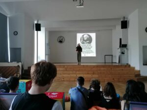 Szeroki kadr ukazujący uczestniczkę wydarzenia stojącą na scenie, przemawiającą do widowni. W tle znany już plakat "Wieczoru Umarłych Poetów". Na pierwszym planie widoczna publiczność siedząca tyłem – kilkanaście osób, w tym chłopcy i dziewczęta. Niektóre osoby siedzą na poduszkach, inne na kolorowych pufach lub podłodze. Panuje przyjazna, nieformalna atmosfera.
