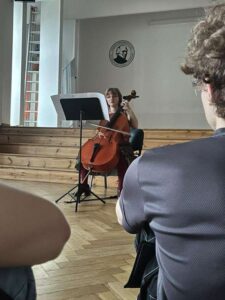 Młoda dziewczyna gra na wiolonczeli. Siedzi na czarnym krześle na tle drewnianej sceny. Ubrana w bordową bluzkę i ciemne spodnie. Przed nią stojak z nutami. Widoczna jest publiczność – młodzi ludzie siedzą na podłodze lub na niskich siedziskach. Ściana w tle biała, z herbem szkoły.