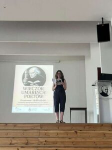 Na scenie stoi młoda kobieta o jasnej karnacji i kręconych brązowych włosach do ramion. Ubrana w ciemny kombinezon bez rękawów i sandały. Trzyma w dłoniach otwartą książkę i recytuje. Za nią na ścianie biały ekran z czarno-białym plakatem. Na plakacie znajduje się rysunkowy portret starszego mężczyzny z surową twarzą, trzymającego świecę i książkę – stylizowany na profesora lub poetę. Pod nim napis:
„WIECZÓR UMARŁYCH POETÓW – 6 czerwca, 19:30–22:00, Liceum im. Stanisława Staszica w Lublinie”.
Sala jasna, minimalistyczna, z drewnianą sceną i podium z herbem szkoły z podobizną Staszica.