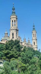 Na zdjęciu widoczna jest boczna fasada tej samej bazyliki (Nuestra Señora del Pilar) z kilku wysokimi, jasnobeżowymi wieżami zakończonymi zielonymi kopułkami i iglicami. Budowla wygląda monumentalnie, z licznymi mniejszymi kopułami w tle pokrytymi kolorowymi dachówkami w geometryczne wzory (w kolorach żółtym, niebieskim, zielonym i białym). Na pierwszym planie widoczne są liczne drzewa o intensywnie zielonych liściach, zasłaniające częściowo dolną część budowli. Niebo jest bezchmurne i intensywnie niebieskie.