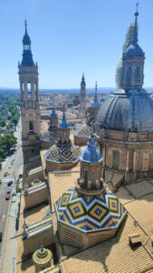 Widok z góry na dach Bazyliki Nuestra Señora del Pilar w Saragossie, w Hiszpanii. Dominują ogromne, kopulaste dachy ozdobione geometrycznymi wzorami z kolorowych dachówek – w kolorach żółtym, niebieskim, zielonym i białym. Dachy mają kształt ośmiokątnych kopuł, a każda z nich wieńczy niebieska, stożkowata wieżyczka. W tle widać kilka strzelistych wież kościoła, a dalej zabudowę miejską z niskimi budynkami i dachami w kolorze cegły. Horyzont sięga aż po odległe wzgórza. Niebo jest jasnoniebieskie, bezchmurne. W lewym dolnym rogu widoczna jest ulica z zaparkowanymi samochodami i linią jezdni prowadzącą do miasta.