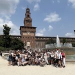 Duża grupa młodzieży pozuje przed monumentalnym zamkiem Castello Sforzesco w Mediolanie. Na środku placu znajduje się okrągła fontanna z wysokim tryskającym strumieniem wody. Za nią rozciąga się ceglana fasada zamku z wieżą zegarową pośrodku. Ceglaste mury ozdobione są białymi detalami i zegarem. W tle po bokach – cylindryczne baszty. Wokół – dużo zieleni i błękitne niebo z białymi chmurami.