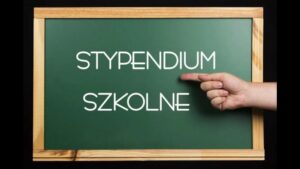 Obraz przedstawia ciemnozieloną szkolną tablicę w drewnianej ramie, umieszczoną na czarnym tle. W centrum tablicy znajduje się biały, prosty, nowoczesny napis pisany wielkimi literami: „STYPENDIUM SZKOLNE”. Dodatkowe elementy: Po prawej stronie obrazu, na wysokości napisu, widoczna jest ludzka dłoń z jasną karnacją, ułożona poziomo z wyprostowanym palcem wskazującym, który wskazuje bezpośrednio na napis „STYPENDIUM”. Tło poza tablicą jest całkowicie czarne, co podkreśla kontrast i skupia uwagę na napisie. Symbolika i kontekst: Obraz w wyraźny sposób wskazuje na temat pomocy finansowej dla uczniów – stypendium szkolne. Użycie szkolnej tablicy i gestu wskazującego ma za zadanie zwrócić uwagę odbiorcy na ważność tematu.