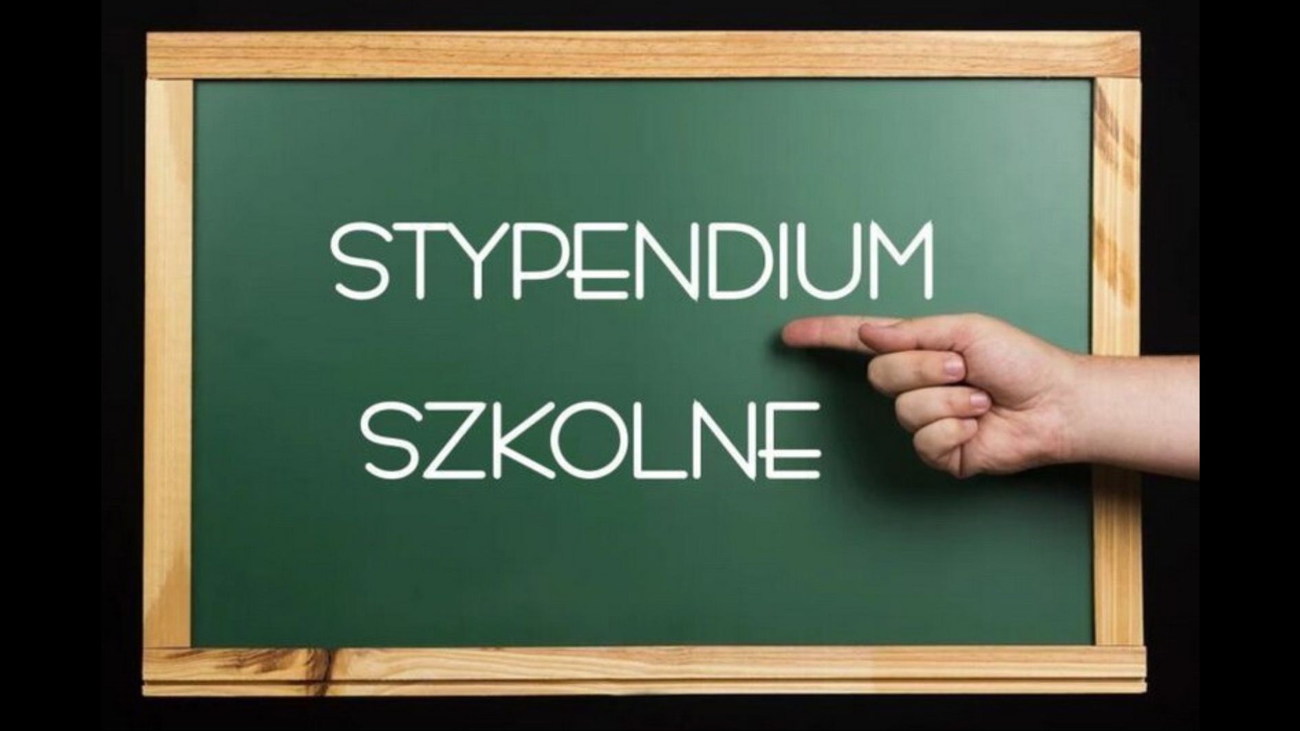 Obraz przedstawia ciemnozieloną szkolną tablicę w drewnianej ramie, umieszczoną na czarnym tle. W centrum tablicy znajduje się biały, prosty, nowoczesny napis pisany wielkimi literami: „STYPENDIUM SZKOLNE”. Dodatkowe elementy: Po prawej stronie obrazu, na wysokości napisu, widoczna jest ludzka dłoń z jasną karnacją, ułożona poziomo z wyprostowanym palcem wskazującym, który wskazuje bezpośrednio na napis „STYPENDIUM”. Tło poza tablicą jest całkowicie czarne, co podkreśla kontrast i skupia uwagę na napisie. Symbolika i kontekst: Obraz w wyraźny sposób wskazuje na temat pomocy finansowej dla uczniów – stypendium szkolne. Użycie szkolnej tablicy i gestu wskazującego ma za zadanie zwrócić uwagę odbiorcy na ważność tematu.