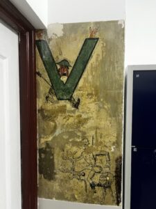 Fragment ściany wewnątrz budynku, z częściowo zachowaną starą, ręcznie malowaną dekoracją. Na tle zniszczonej, popękanej i miejscami odpadającej żółto-brązowej farby widoczna jest wielka, ciemnozielona litera „V”. Z wnętrza litery wychyla się postać narysowanego mężczyzny w zielonym kapeluszu z rondem, mającego wąsy i wyciągnięte w górę ramiona zakończone dłońmi w geście zwycięstwa (palce w kształcie litery „V”). Niżej, na tym samym fragmencie ściany, widać czarny szkic wykonany cienką kreską – przedstawia on jeźdźca siedzącego na reniferze lub jeleniu, trzymającego go za poroże. Zwierzę i jeździec są schematyczni, bez wypełnienia kolorem. Ściana znajduje się w rogu między drzwiami w drewnianej, ciemnoczerwonej ramie a szafką o niebieskich drzwiczkach.