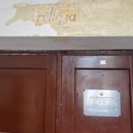 Górna część dużych, podwójnych drzwi z ciemnobrązowego drewna. Nad nimi widoczny fragment ściany, na którym spod odłupanej warstwy farby odsłonił się stary napis w języku polskim: „religia” – litery są lekko pochylone, w kolorze szaroczarnym na żółtawym tle. Farba wokół napisu jest w dużej części zdarta, pozostawiając nieregularne krawędzie. Na prawym skrzydle drzwi znajduje się prostokątna, metalowa tabliczka pamiątkowa w kolorze srebrnym z czarnym napisem: „SALA MATEMATYCZNA” wraz z informacjami o patronie i dacie. Tabliczka jest przykręcona czterema śrubami, po jednej w każdym rogu. Po lewej stronie na drzwiach widoczna jest niewielka kłódka lub element zamka.